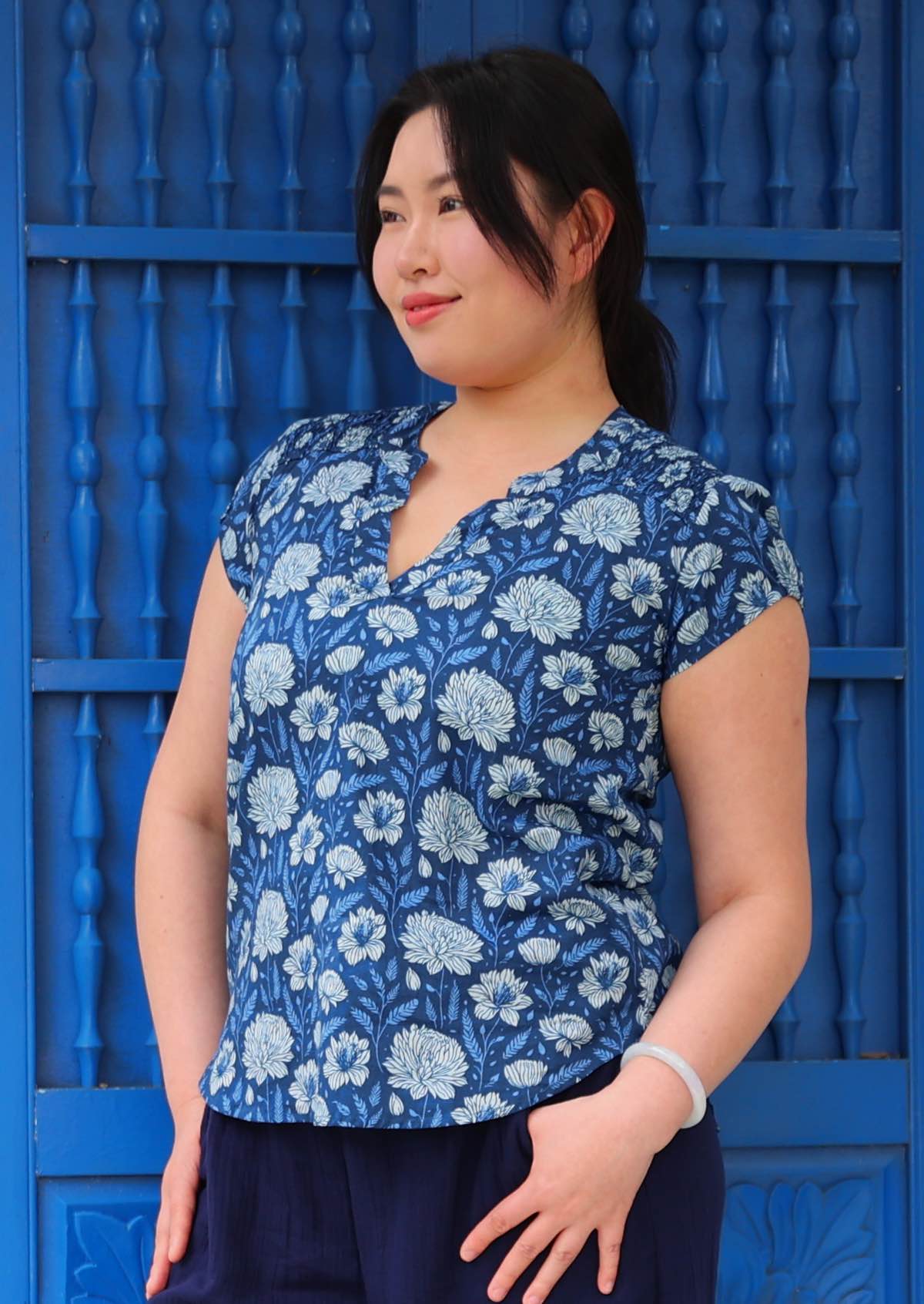 Woman with black hair wearing a blue cotton top with cp sleeves in light blue and white floral print, in front of blue door
