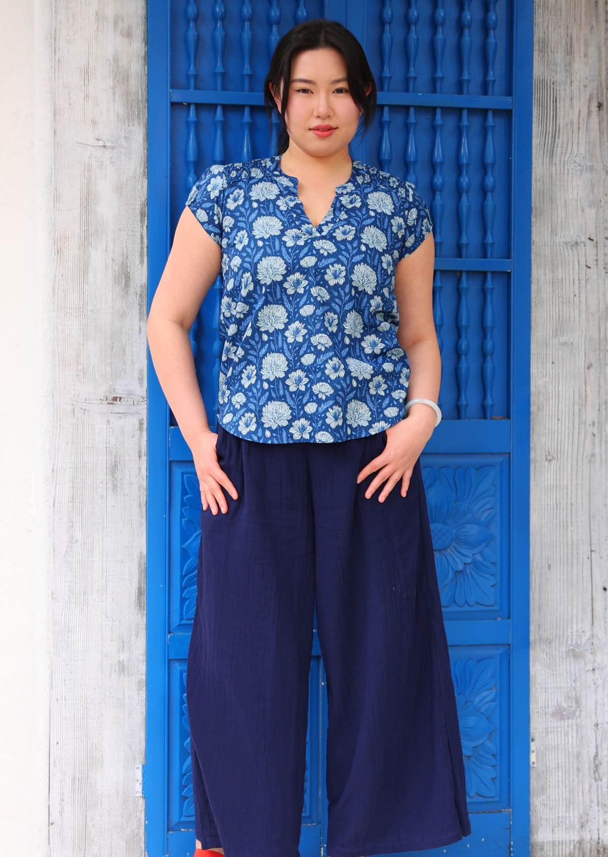 Woman with black hair wearing a blue cotton top with light blue and white floral print, with mandarin collar, in front of blue door
