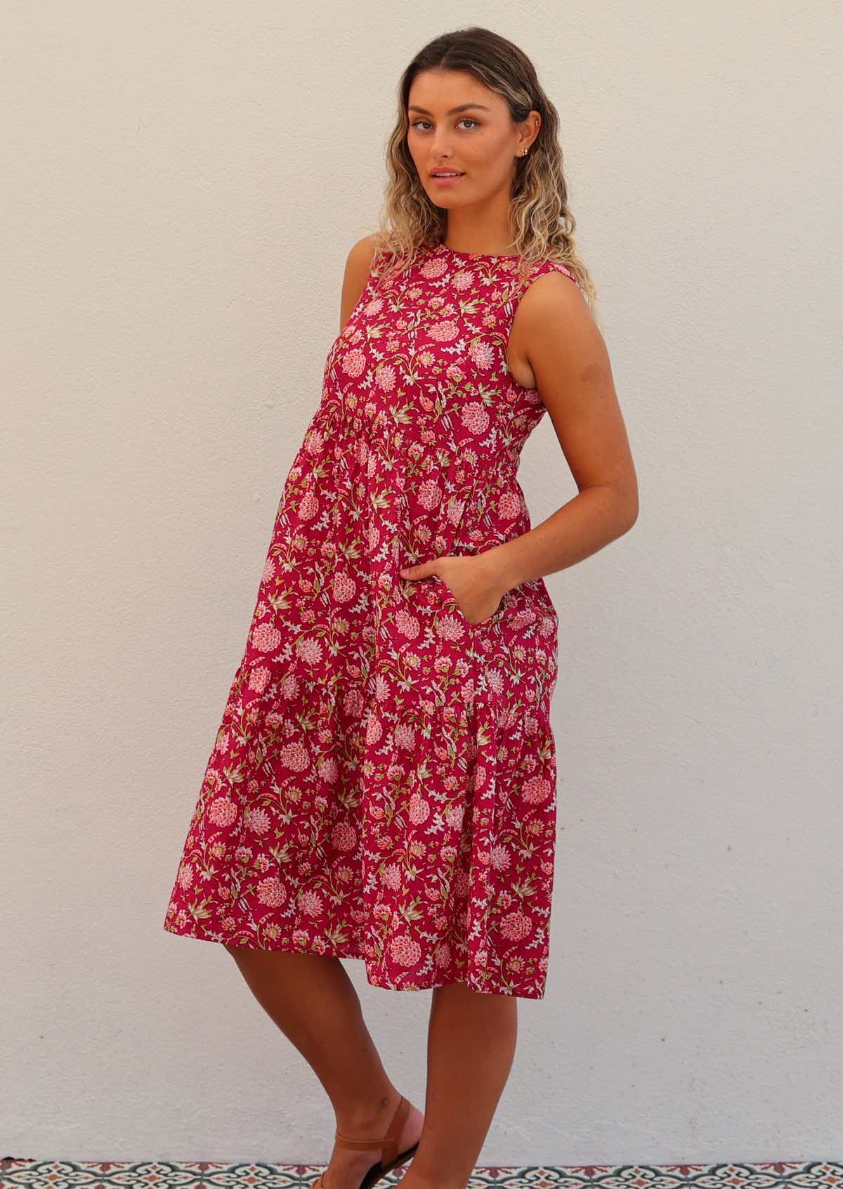 Woman wears 100% cotton sleeveless midi length dress in floral print on a cranberry base with hand in pocket