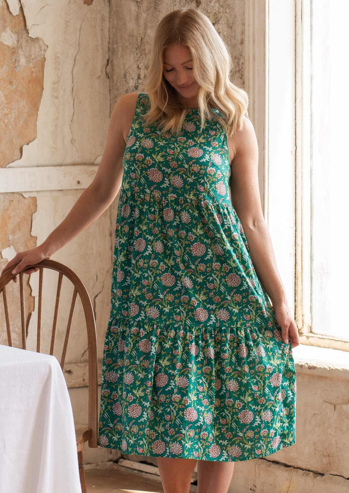 Woman holds skirt of cotton midi length sleeveless dress out, in floral print with jade green base