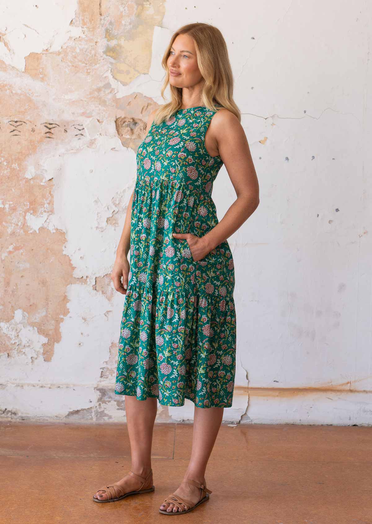 Model stands sideways in cotton sleeveless midi length dress in floral print with hand in hidden side pocekt