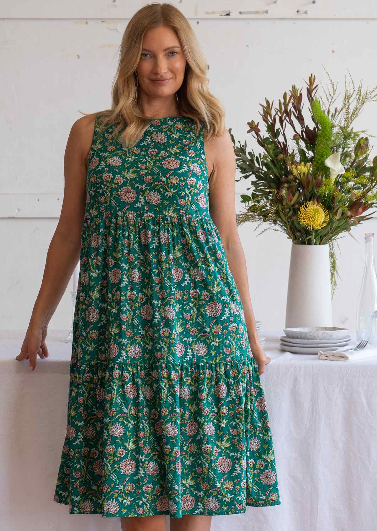 Woman stands in front of table in jade green based floral print sleeveless midi length dress with high round neckline