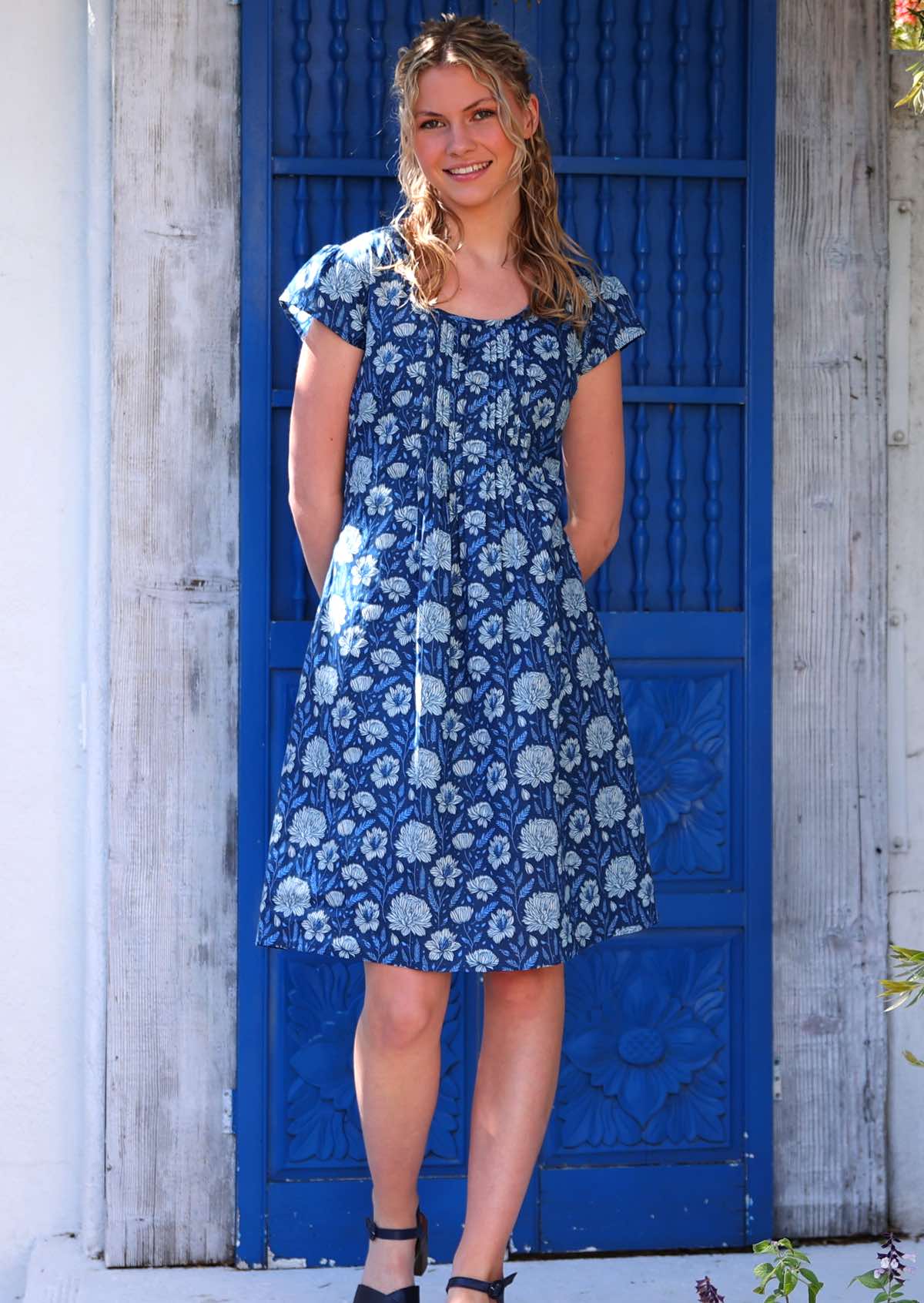 Woman with blonde hair wearing a blue cotton knee length dress with round neckline in light blue and white floral design, in front of blue door
