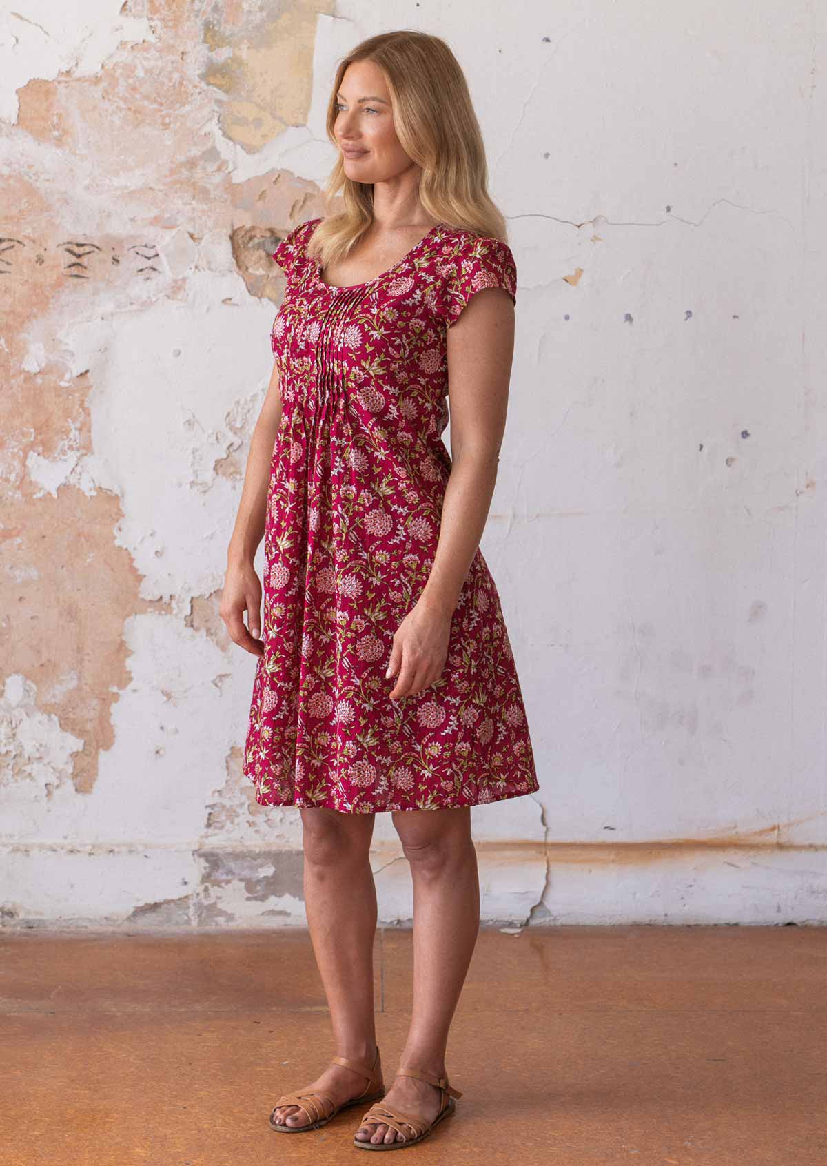 Woman stands sideways in above knee cotton dress with small pleats across bodice, in a red based floral print