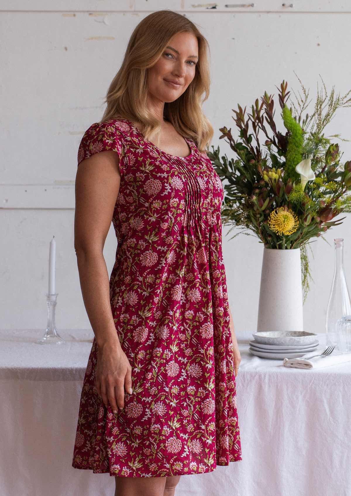 Woman wears cotton cap sleeve above knee dress with round neckline and small pleats across the bodice, in a red based floral print