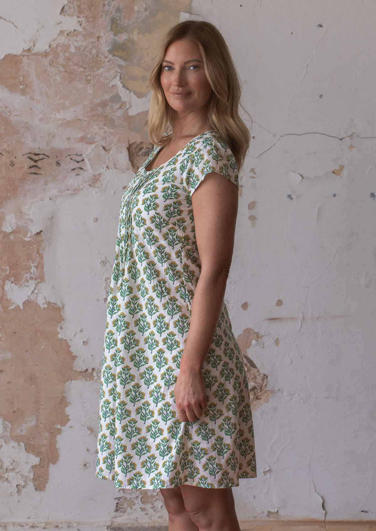 Model stands in cotton above knee dress with cap sleeves, round neckline and pin tucks across bodice