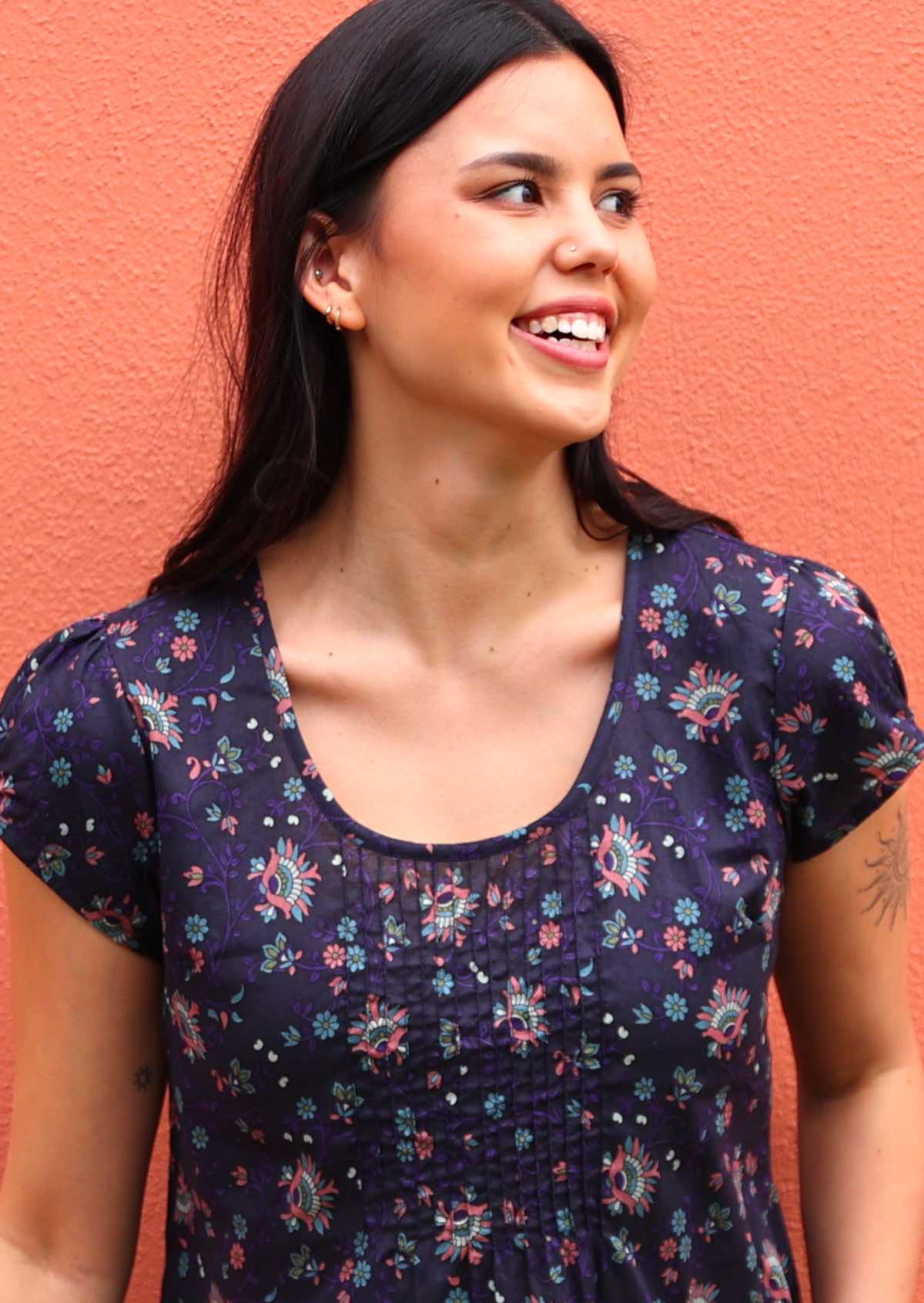 Close up of model posing in cotton cap sleeve top with deep round neckline and small pleats across bodice, in dark based floral print