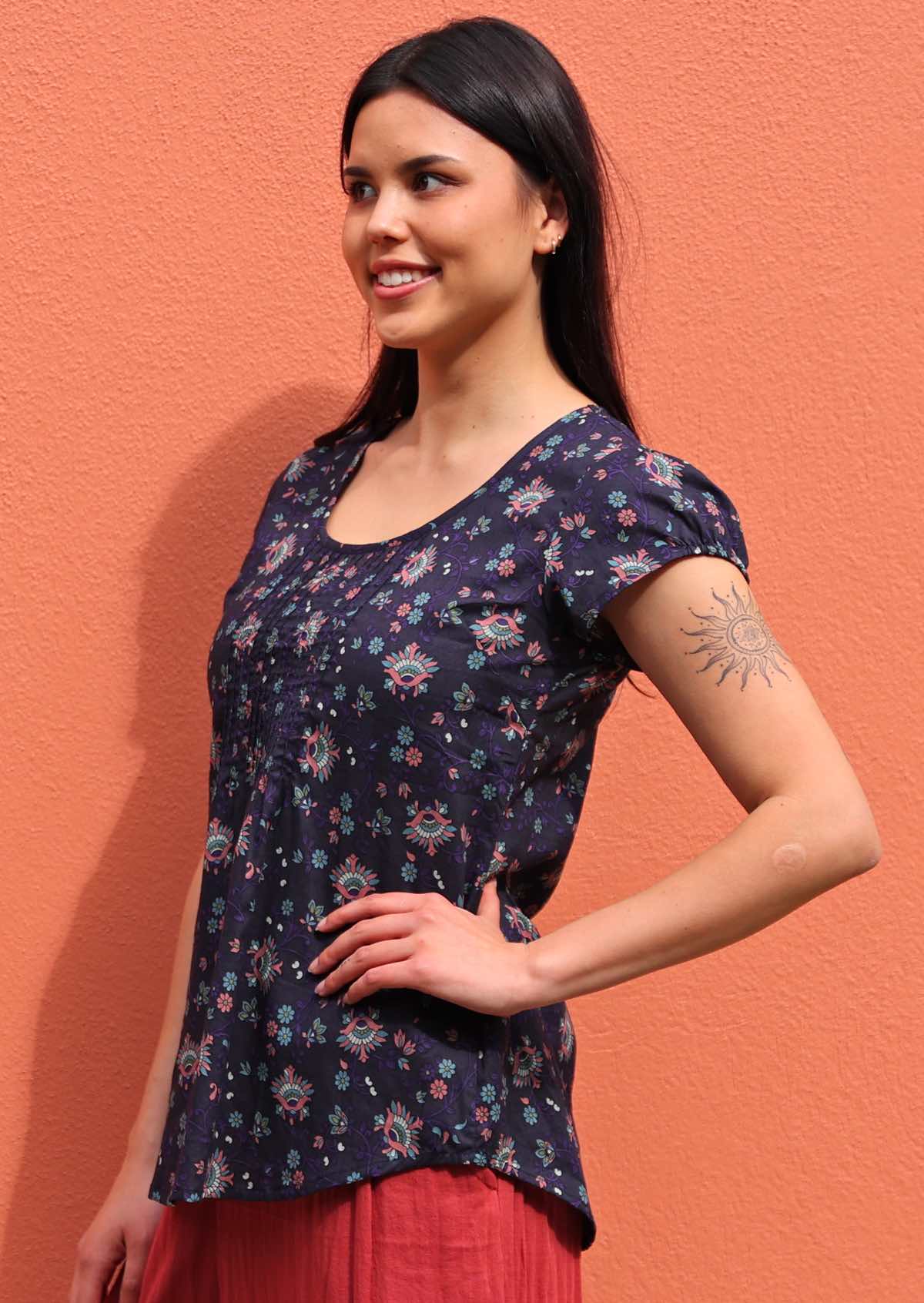 Model posses to the side in 100% cotton cap sleeve floral top with deep round neckline and small pleats across bodice