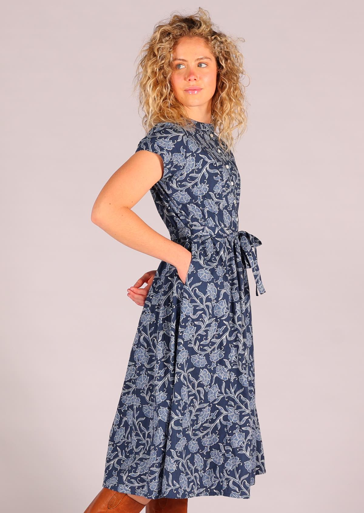 Model standing side on with hand in pocket 100 percent cotton retro button down shirt dress with pockets and waist tie in floral and navy blue background