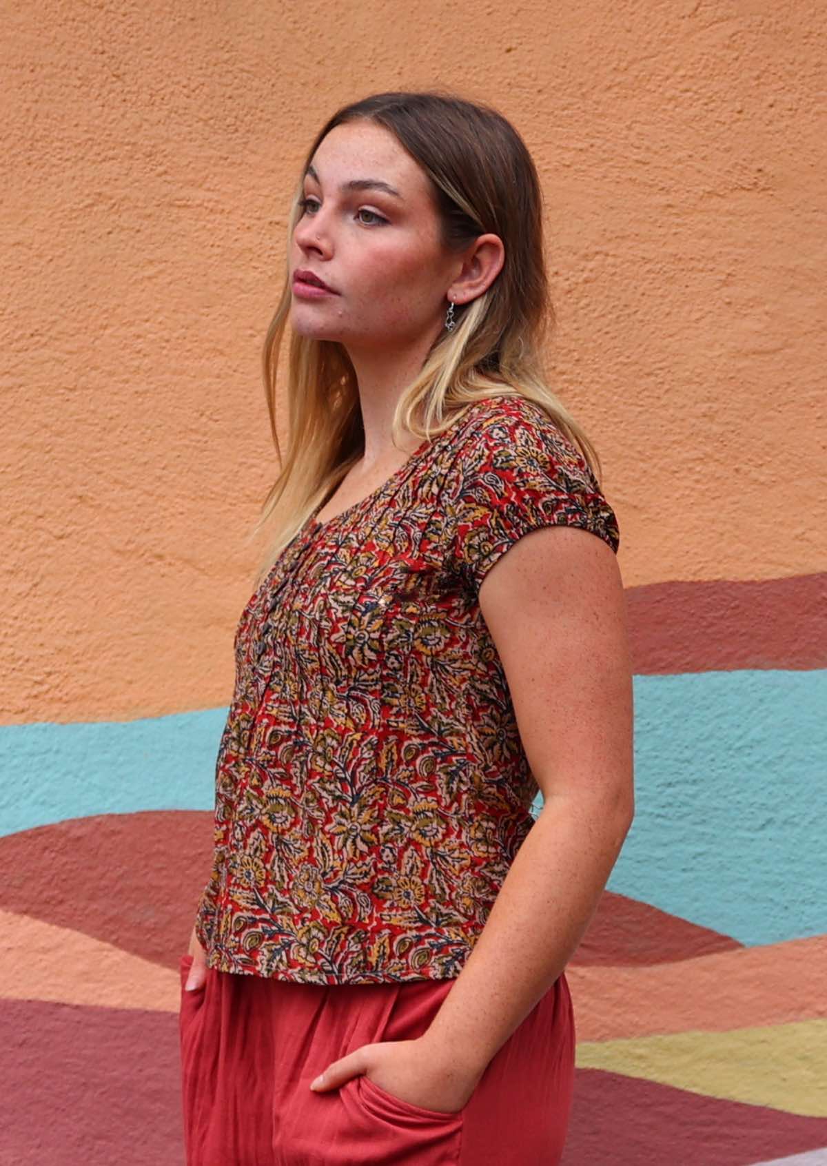 Model side on with hands in pant pockets wearing 100 percent cotton Indian block print blouse florals on a red base with buttons